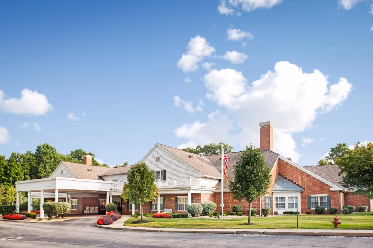 Front exterior of a brick and siding senior living building with a covered entrance, landscaping, and an American flag under a blue sky.