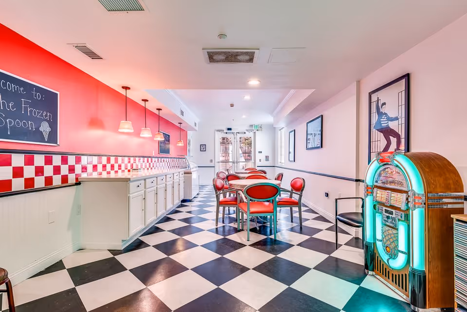 Interior of a retro-style ice cream parlor with black and white checkered floor, red and white tiled walls, red chairs around tables, a jukebox on the right, and a chalkboard sign that reads 'Welcome to the Frozen Spoon'.