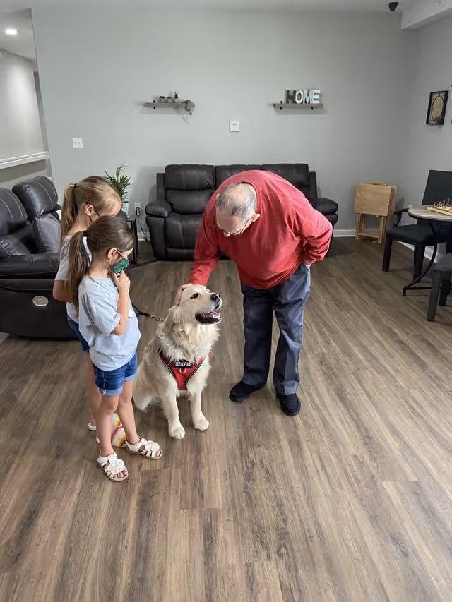 An elderly man in a red sweater is petting a golden retriever wearing a red service dog vest. Two young girls stand nearby, one holding the dog's leash. They are in a living room with wooden floors, dark leather sofas, a small table with chairs, and wall shelves with decorative items.