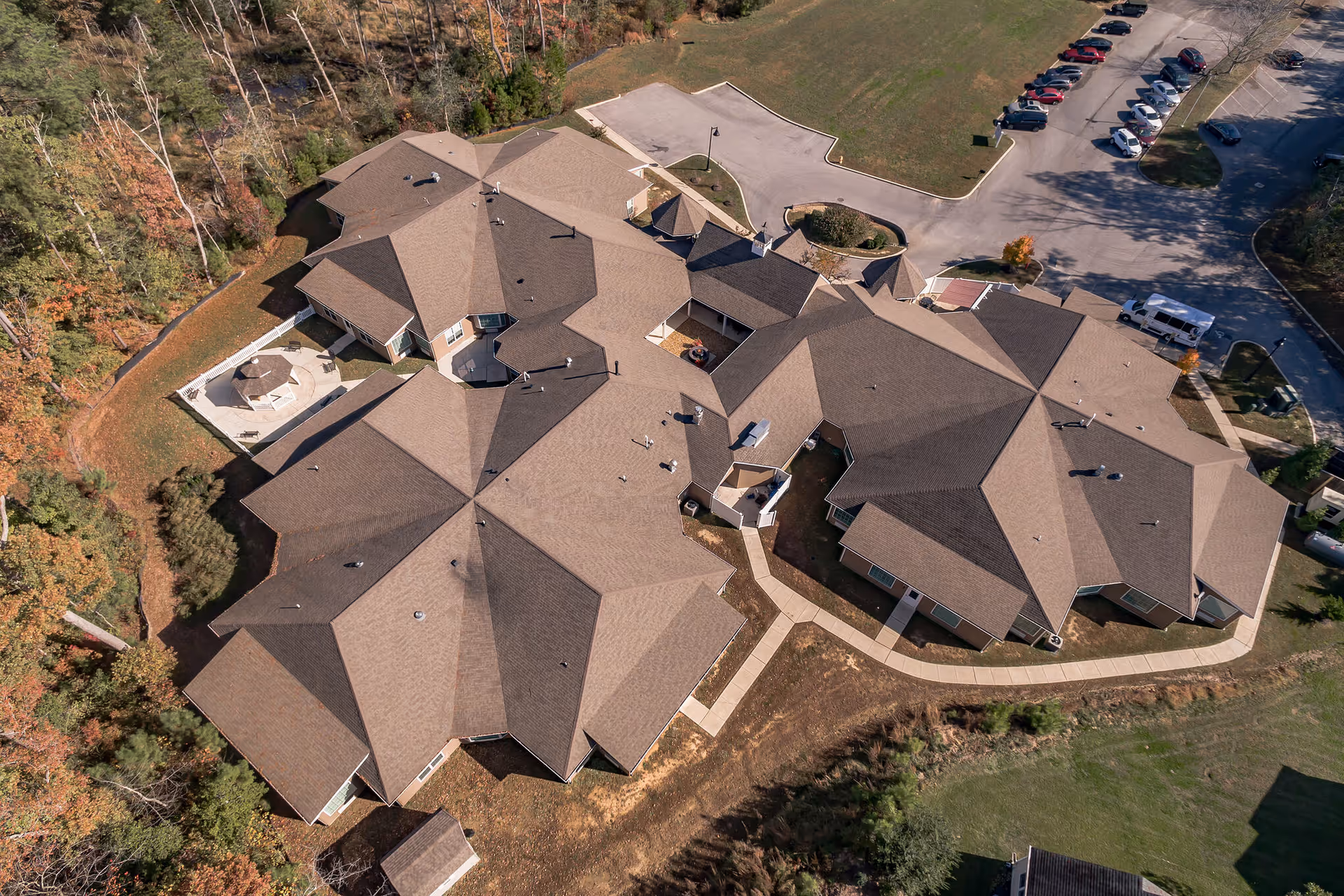 Aerial view of a large senior living facility building with multiple roof sections, surrounded by trees and a parking lot with several cars.