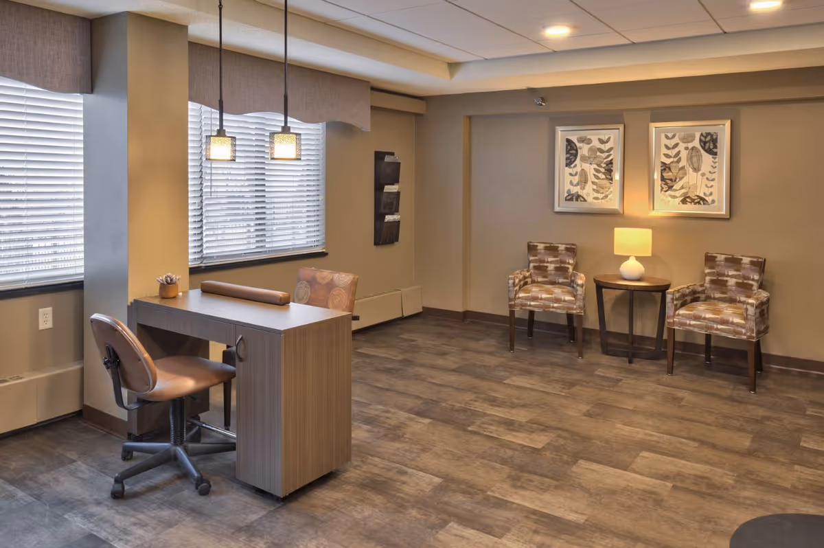 A well-lit interior room with two patterned armchairs and a small round table with a lamp between them against a beige wall. Two framed botanical prints hang above the chairs. To the left, there is a wooden desk with a brown office chair and another patterned chair, positioned near two large windows with blinds and pendant lights hanging above the desk. The floor has a wood-like finish.