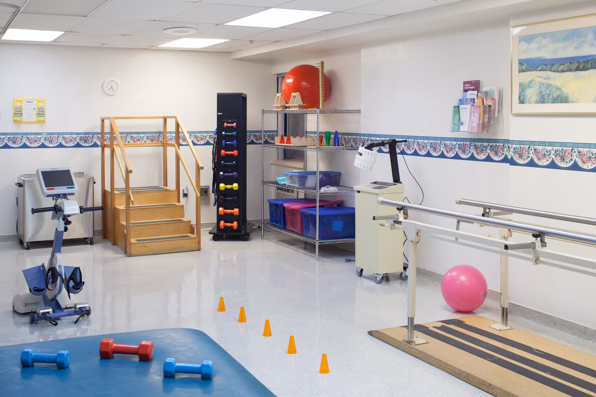A physical therapy or rehabilitation room with exercise equipment including a stationary bike, small dumbbells, a set of stairs with handrails, parallel bars for walking exercises, a pink exercise ball, and shelves holding various therapy tools and cones. The room has white walls with a floral border and a framed landscape painting.