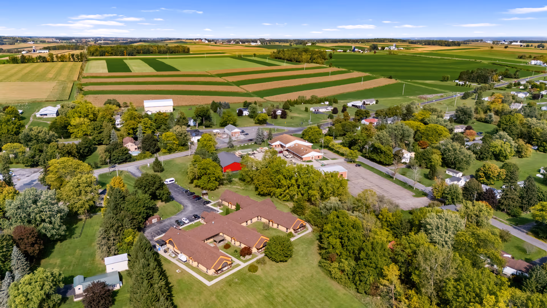 Aerial view of Clinton Crest Manor and surrounding rural area with green fields, trees, and scattered houses under a clear blue sky.