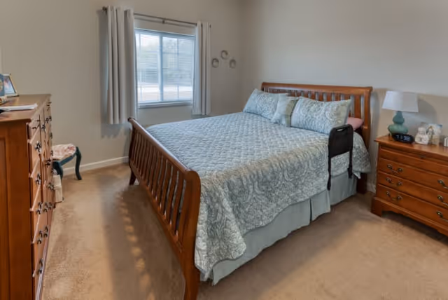 A cozy bedroom with a wooden bed frame and a neatly made bed with a light blue patterned quilt and matching pillows. There is a wooden dresser on the left side and a wooden nightstand with a lamp, clock, and decorative items on the right. A window with light-colored curtains lets in natural light, and the walls are painted a soft beige color.