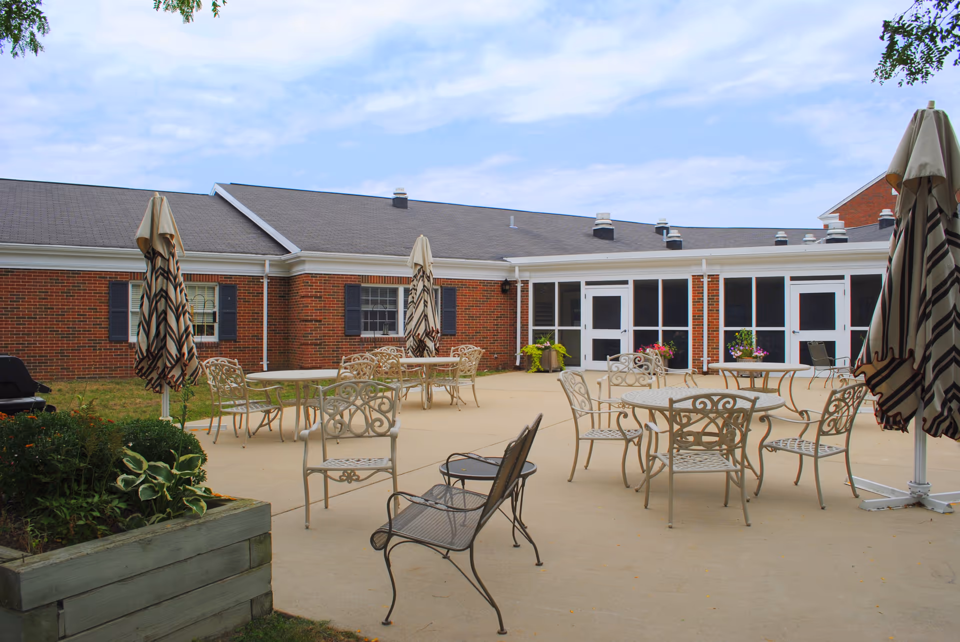 Outdoor paved patio with metal tables, chairs, and closed umbrellas in front of a single-story brick building.