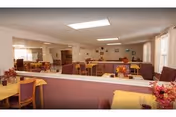 Communal dining area with multiple tables and chairs, yellow tablecloths, and floral centerpieces in a senior facility.
