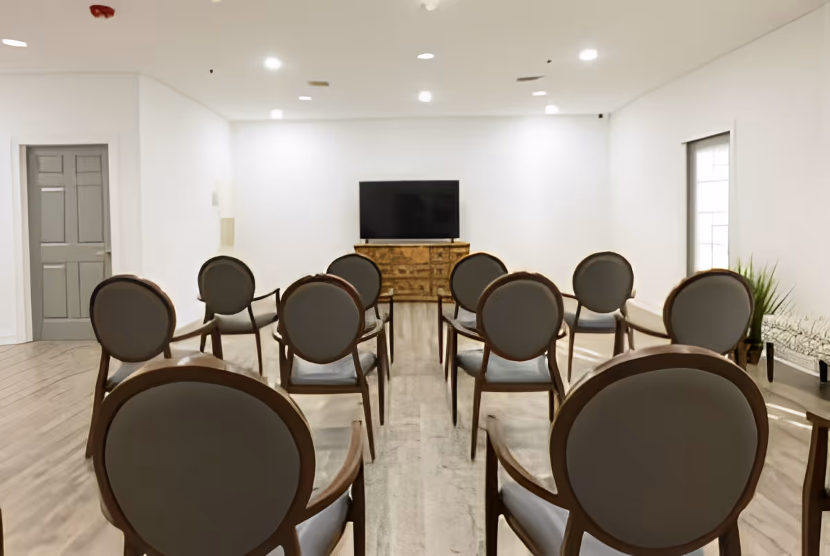 A small meeting or presentation room with rows of wooden chairs facing a flat-screen TV mounted on a wooden cabinet. The room has white walls, a wooden floor, a closed gray door on the left, and a window on the right with a small plant on a table beneath it.