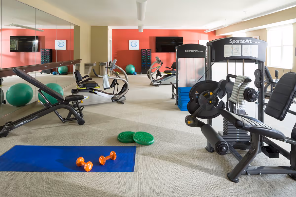A bright fitness room with various exercise equipment including weight machines, stationary bikes, a bench, exercise balls, dumbbells on a blue mat, and balance discs. The room has a large mirror on one wall, a red accent wall with a mounted TV, and windows letting in natural light.