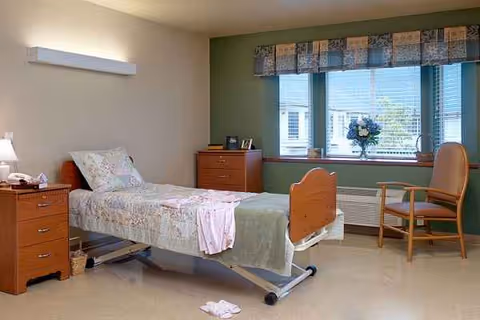 Hospital-style patient bedroom with adjustable bed, nightstands, dresser, window view, and chair.