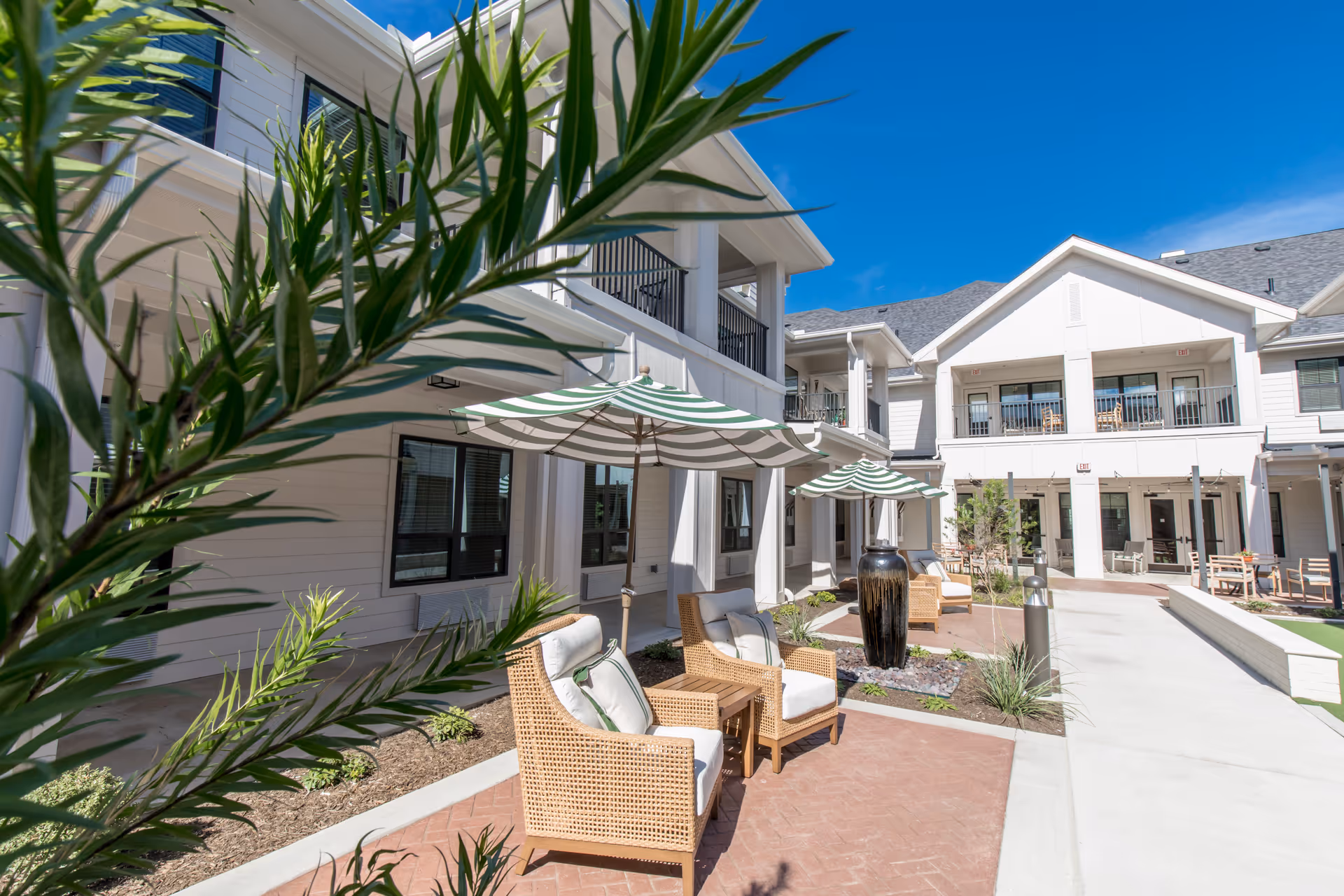 Outdoor courtyard area of The Blake at Waco senior living facility featuring wicker chairs with cushions, striped green and white umbrellas, potted plants, and a two-story building with balconies under a clear blue sky.