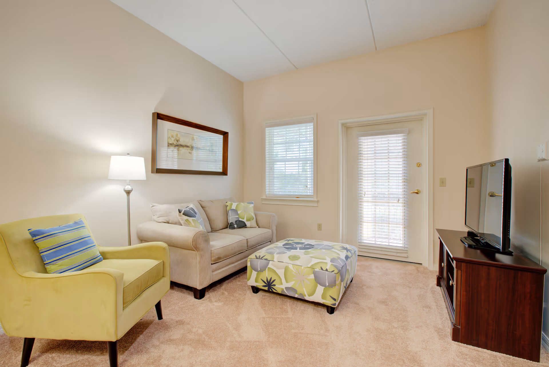 A cozy living room with a beige sofa adorned with patterned cushions, a green armchair with a striped pillow, a floral patterned ottoman, a floor lamp, a framed picture on the wall, a window with blinds, and a door with blinds. A flat-screen TV sits on a wooden TV stand on the right side.