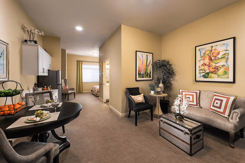 Furnished assisted-living suite showing a living area with a sofa, chairs and trunk coffee table, a dining table and kitchenette, with a bedroom visible down the hallway.