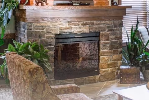 Cozy living room area featuring a stone fireplace with a wooden mantle, surrounded by green plants and a patterned upholstered chair. The room has a warm and inviting atmosphere with natural light coming through window blinds.