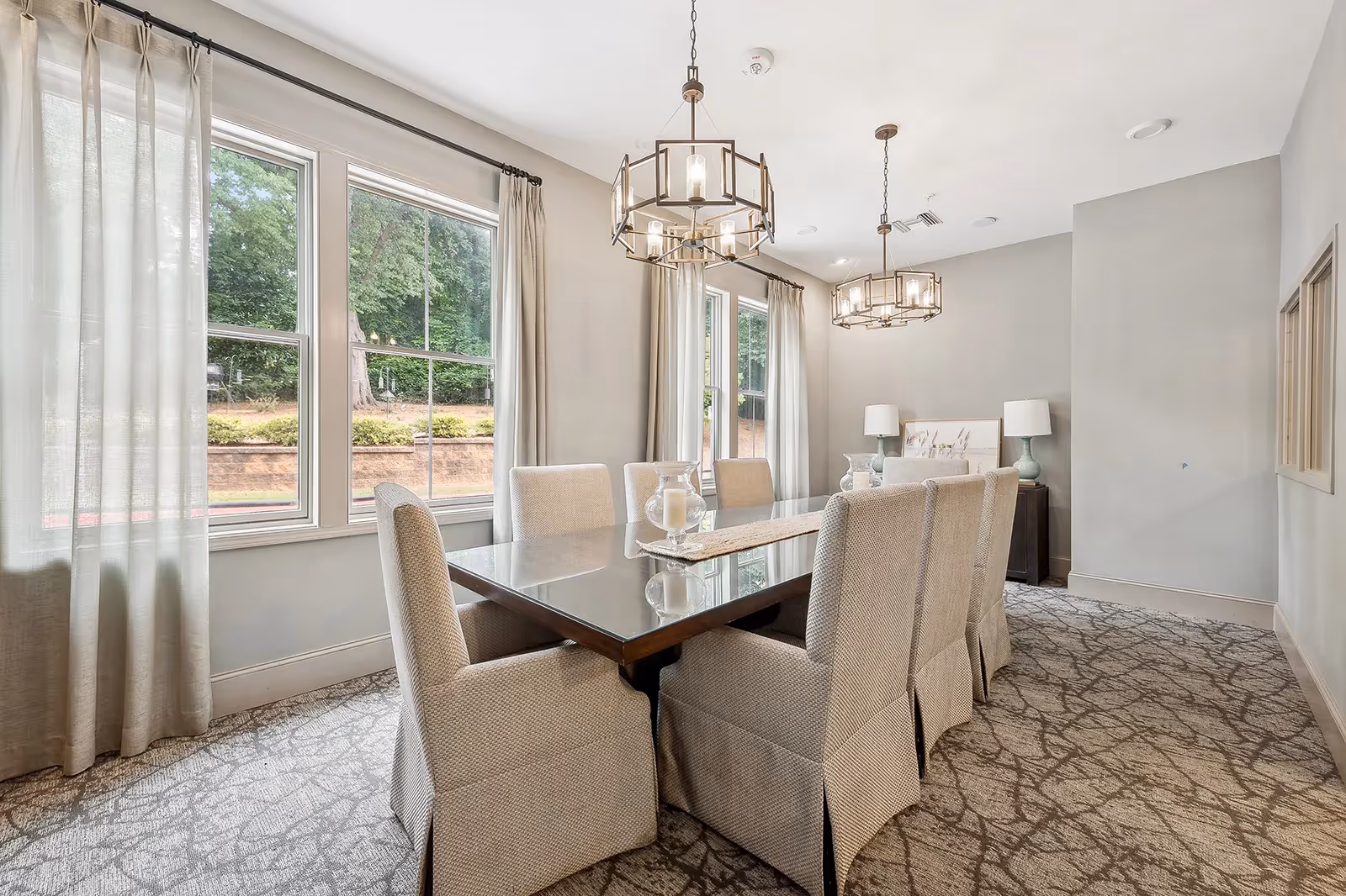 Bright, modern dining room with a long glass-top table, upholstered chairs, two chandeliers, and large windows with curtains.