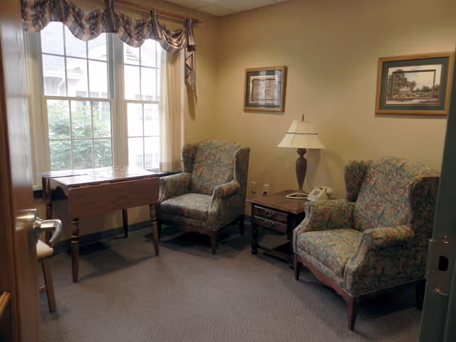A small sitting room with two patterned armchairs, a side table and lamp, a folding table by a window with curtains.
