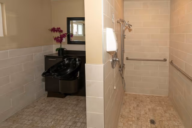 A tiled bathroom area with a black hair washing sink and cabinet on the left side, decorated with a purple orchid plant and a mirror above. On the right side, there is a walk-in shower with beige tiles, grab bars, a showerhead, and a soap dispenser mounted on the wall.