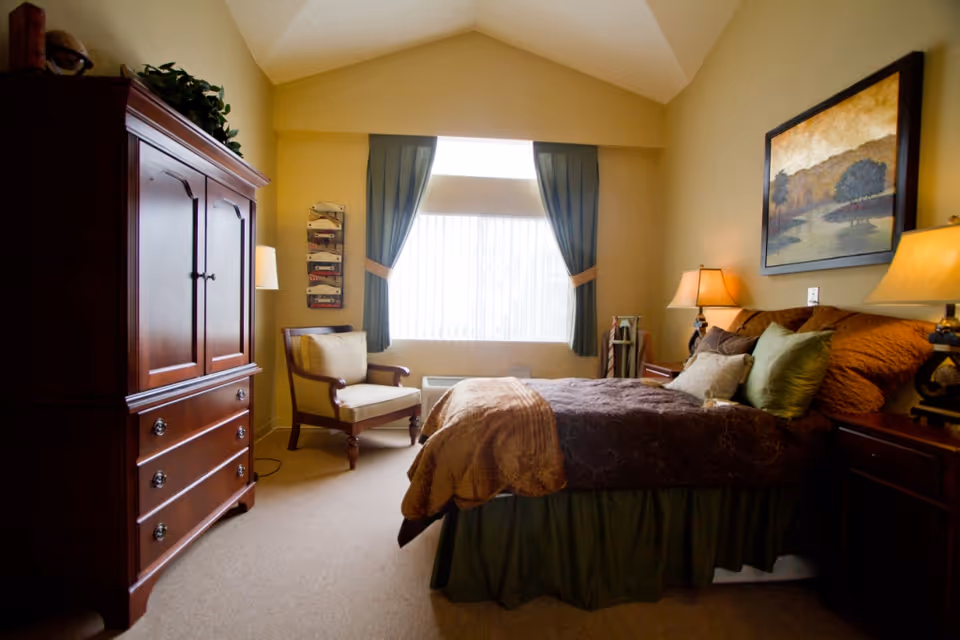 A cozy bedroom with a large bed covered in brown and green bedding, two bedside tables with lamps, a wooden armoire, a cushioned armchair near a window with green curtains, and a framed landscape painting on the wall.