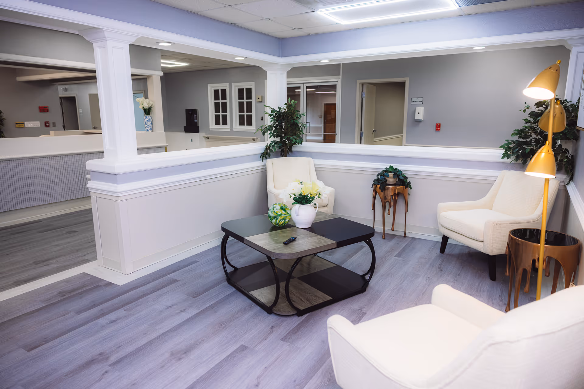 A bright and clean sitting area in a healthcare facility featuring three cream-colored armchairs arranged around a black and wood coffee table with decorative items including a white vase with flowers and a green decorative ball. The space has light gray walls, wood flooring, white trim, a yellow floor lamp, and some green plants. In the background, there is a reception desk and hallway.