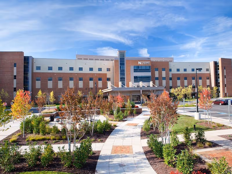 Front exterior view of a large multi-story brick and glass building with a landscaped garden and paved walkway leading to the entrance under a clear blue sky.
