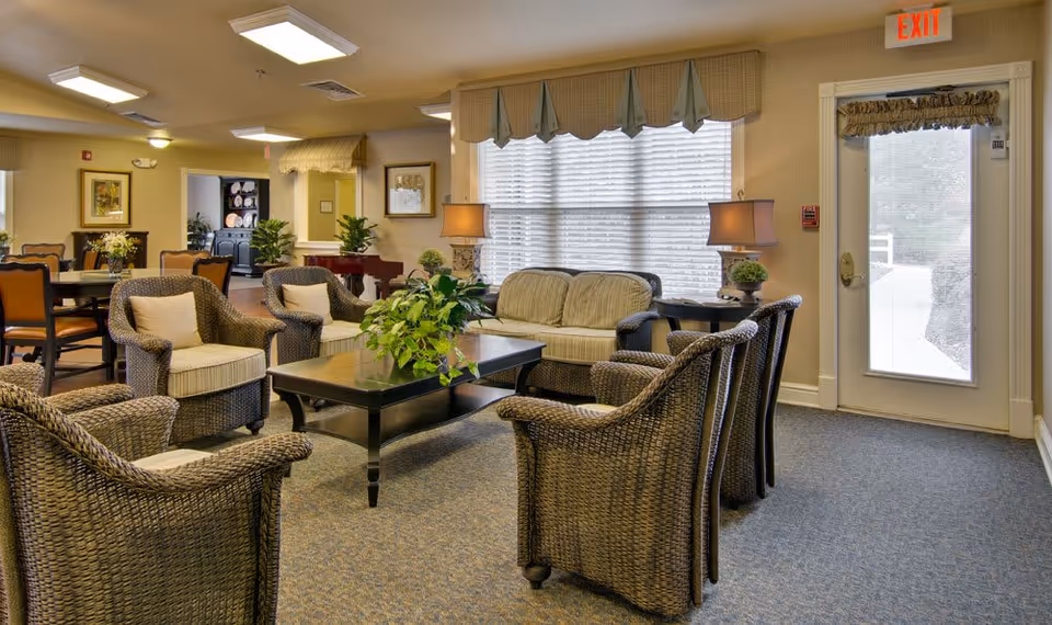 A cozy living room area in a senior living facility with wicker armchairs and a cushioned sofa arranged around a black coffee table with a green plant. The room has large windows with blinds and valances, two table lamps, and a door with an exit sign above it. In the background, there is a dining area with tables and chairs, framed artwork on the walls, and additional plants.