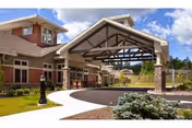 Exterior view of a senior living facility named RiverMead featuring a covered entrance with a peaked roof supported by stone pillars, a curved driveway, landscaped greenery, and a partly cloudy blue sky.