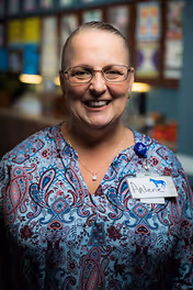 A smiling woman wearing glasses and a patterned blouse with a name tag that reads 'Arlene' standing indoors with colorful artwork displayed on the wall behind her.