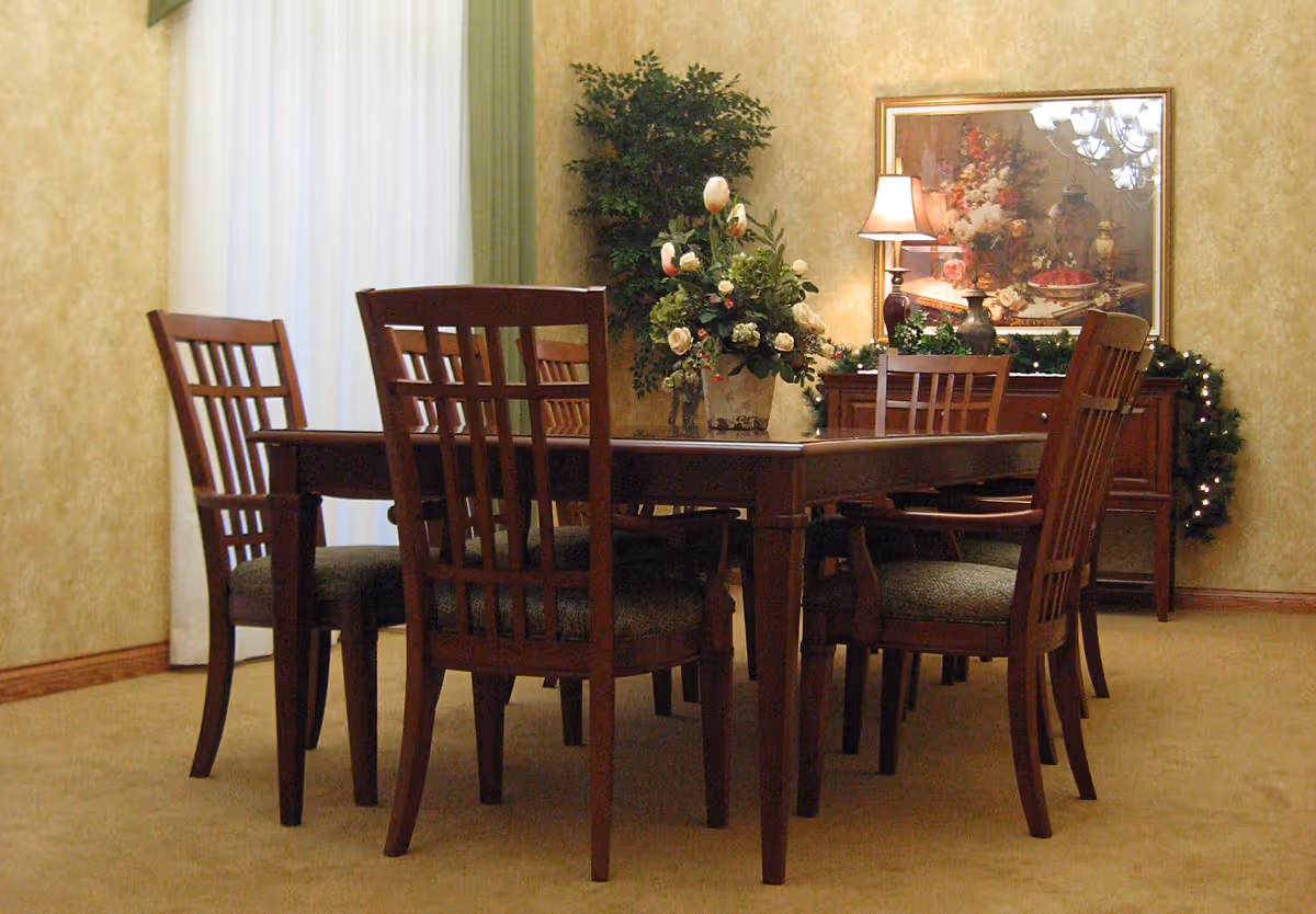 A dining room with a wooden dining table surrounded by six wooden chairs with cushioned seats. On the table is a floral arrangement with white and pink flowers. In the background, there is a sideboard with a table lamp, decorative items, and a framed floral painting on the wall. The room has beige walls, a carpeted floor, and a window with white curtains and green drapes.