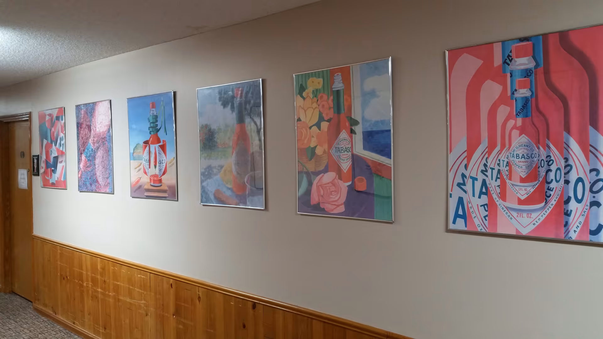 A hallway with six framed colorful paintings of Tabasco sauce bottles hanging on a beige wall above wooden paneling. There is a closed wooden door at the end of the hallway.