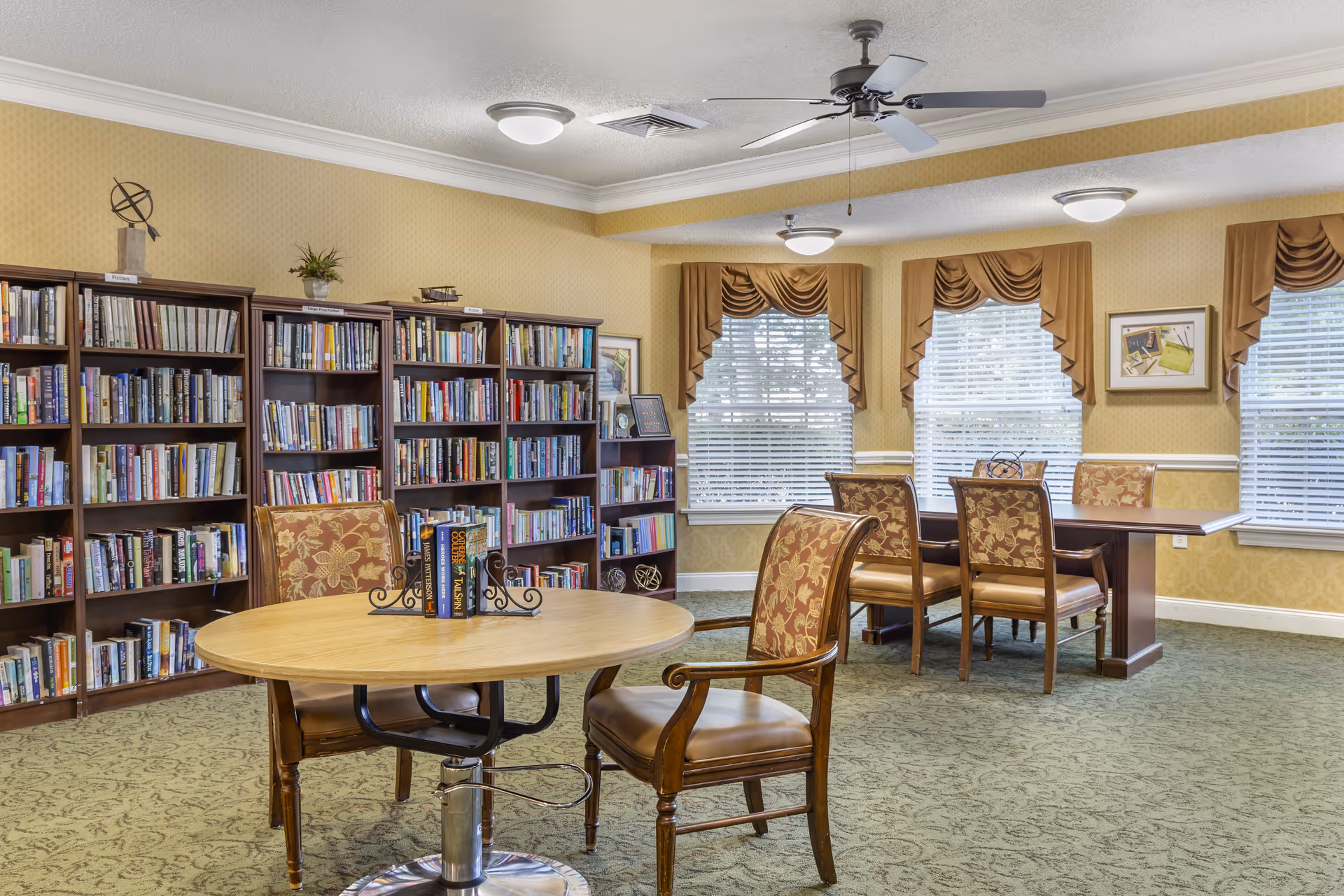 Communal library-style room with bookshelves, round and rectangular tables, and upholstered chairs by bright windows.