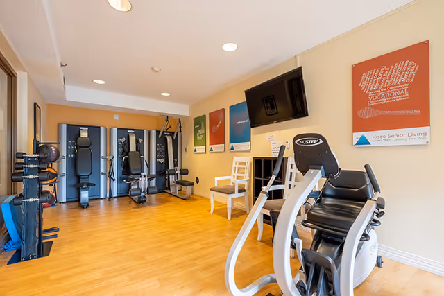 A senior living facility exercise room with various fitness equipment including a NuStep recumbent cross trainer, weight machines, and a rack of dumbbells. The room has light wood flooring, beige walls, and several colorful motivational posters on the wall. A flat-screen TV is mounted on the wall above two chairs.