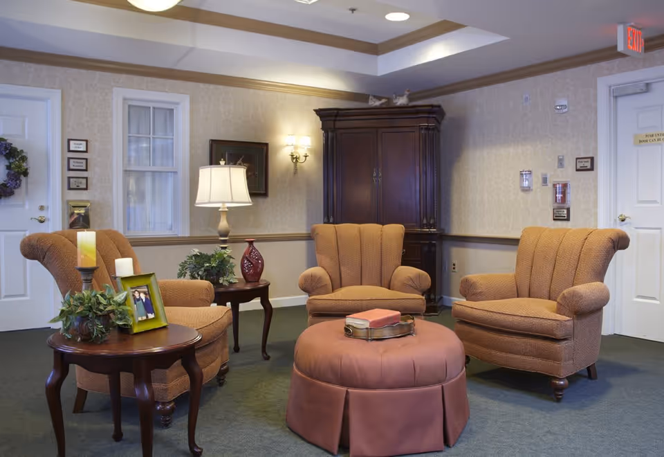 A cozy sitting area in a senior living facility with three upholstered armchairs arranged around a round ottoman. There are two wooden side tables with decorative items including a lamp, framed photo, plants, and candles. The room has beige wallpaper, green carpet, and a wooden cabinet against the wall. Two white doors and a small window are visible in the background.