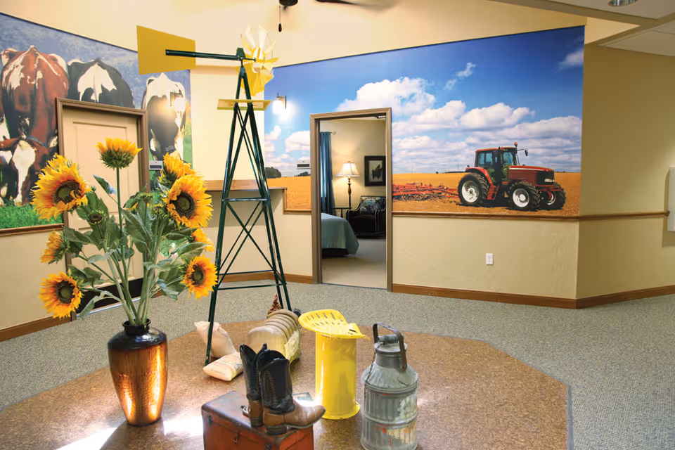 Interior common area with a vase of sunflowers, decorative windmill and farm-themed mural showing a tractor, and an open doorway to a bedroom.