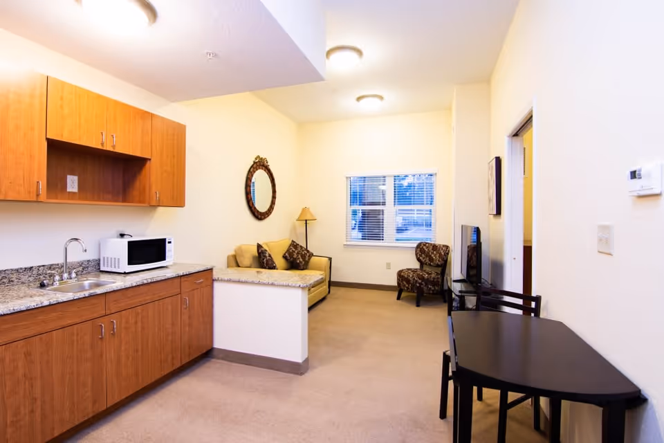 Interior view of a senior living facility room at Royal Dalton House featuring a small kitchenette with wooden cabinets, a microwave, and a sink on the left. The room also includes a beige loveseat with decorative pillows, a standing lamp, a round mirror on the wall, a patterned armchair near a window with blinds, a flat-screen TV on a stand, and a black dining table with chairs on the right.