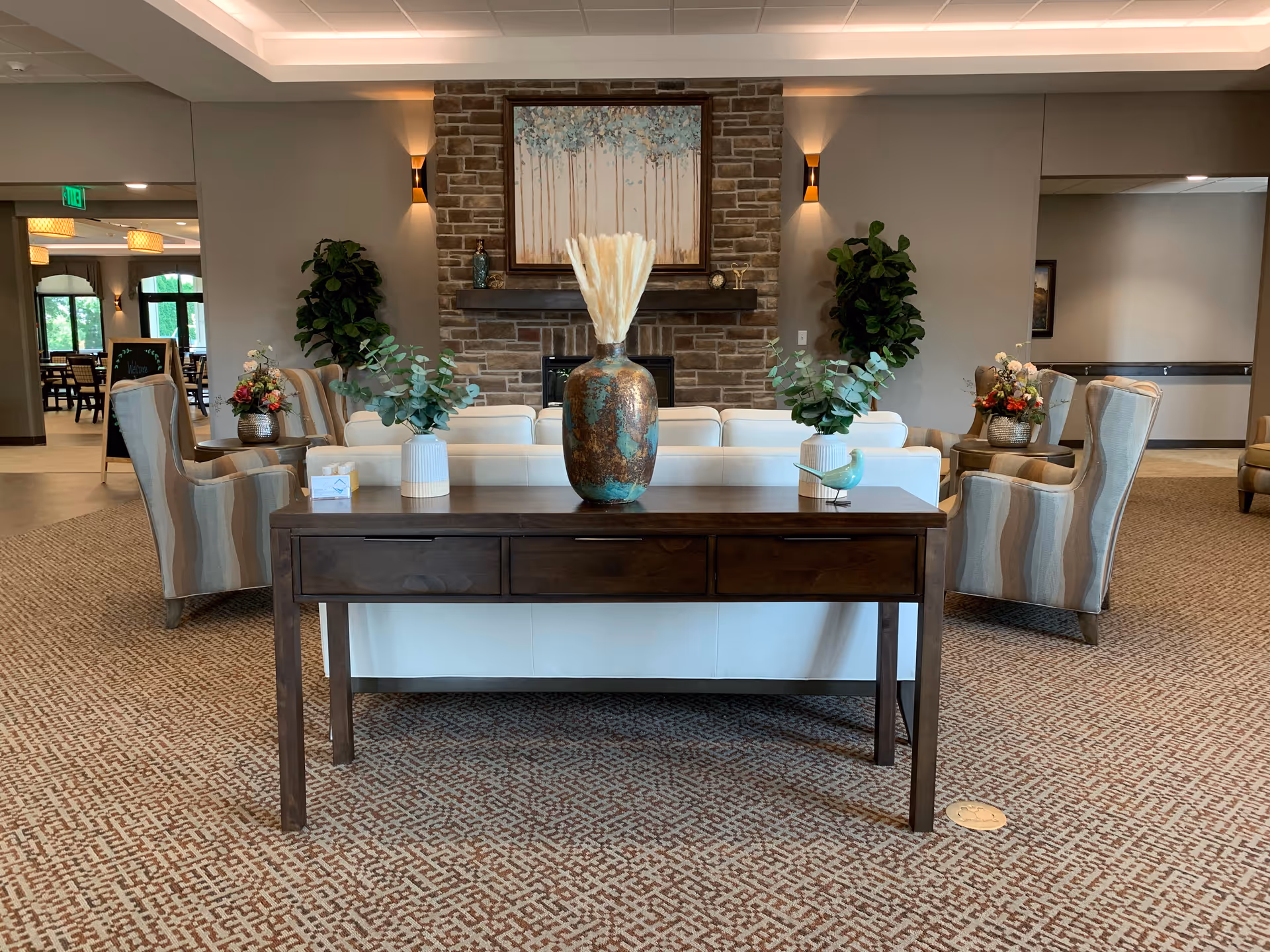 A cozy living room area in CopperStone Assisted Living featuring a dark wooden console table with decorative vases and greenery on top. Behind the table is a white sofa, two striped armchairs, and a stone fireplace with a painting of trees above it. The room has warm lighting and carpeted flooring, with additional seating and plants visible in the background.