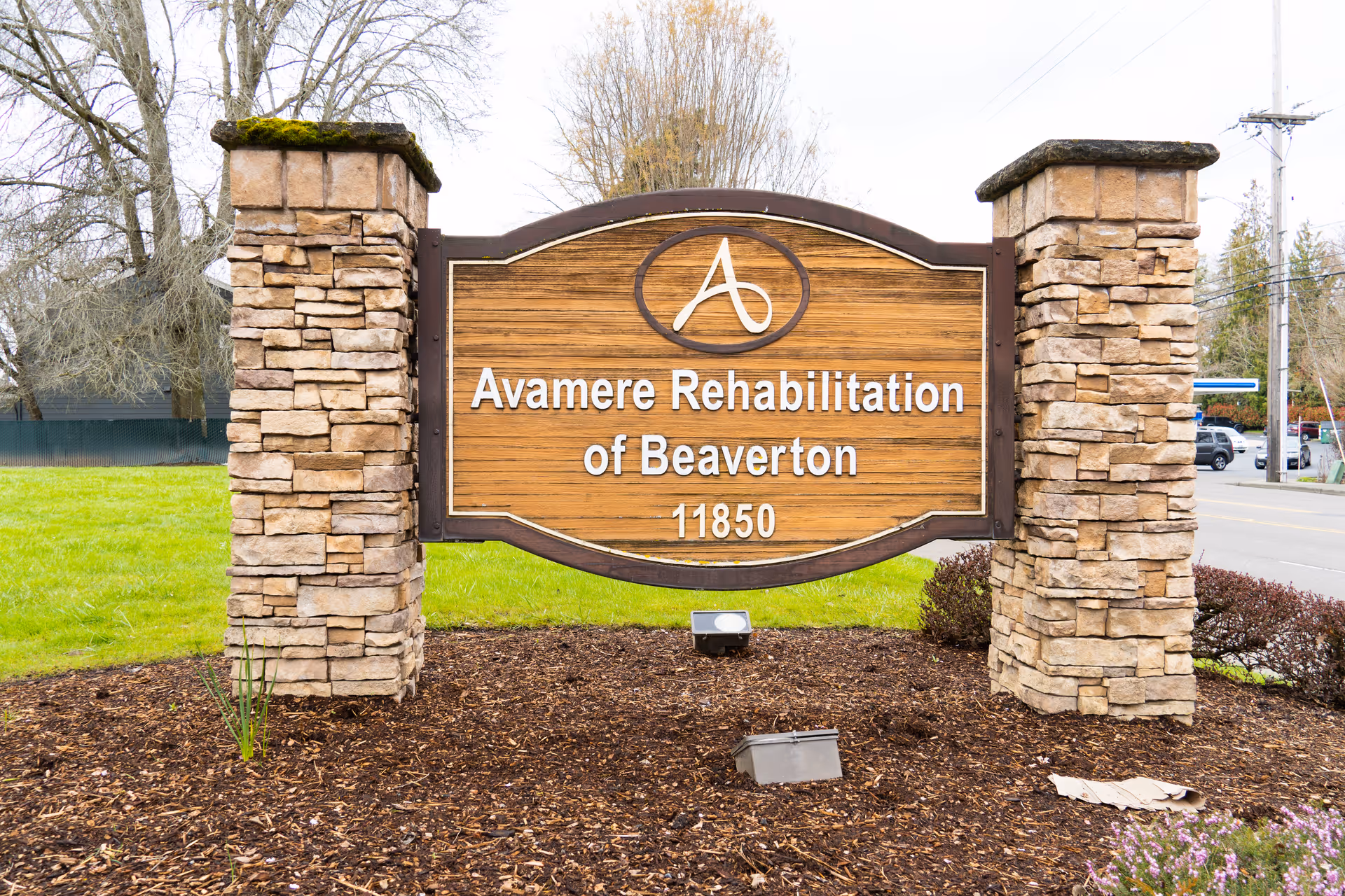 A wooden sign for Avamere Rehabilitation of Beaverton mounted between two stone pillars, with grass and trees in the background and a road to the right.