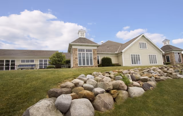 Exterior view of a light-colored retirement village building with large windows, stone accents, and a sloped roof under a partly cloudy sky. The foreground features a grassy lawn and a rock garden.