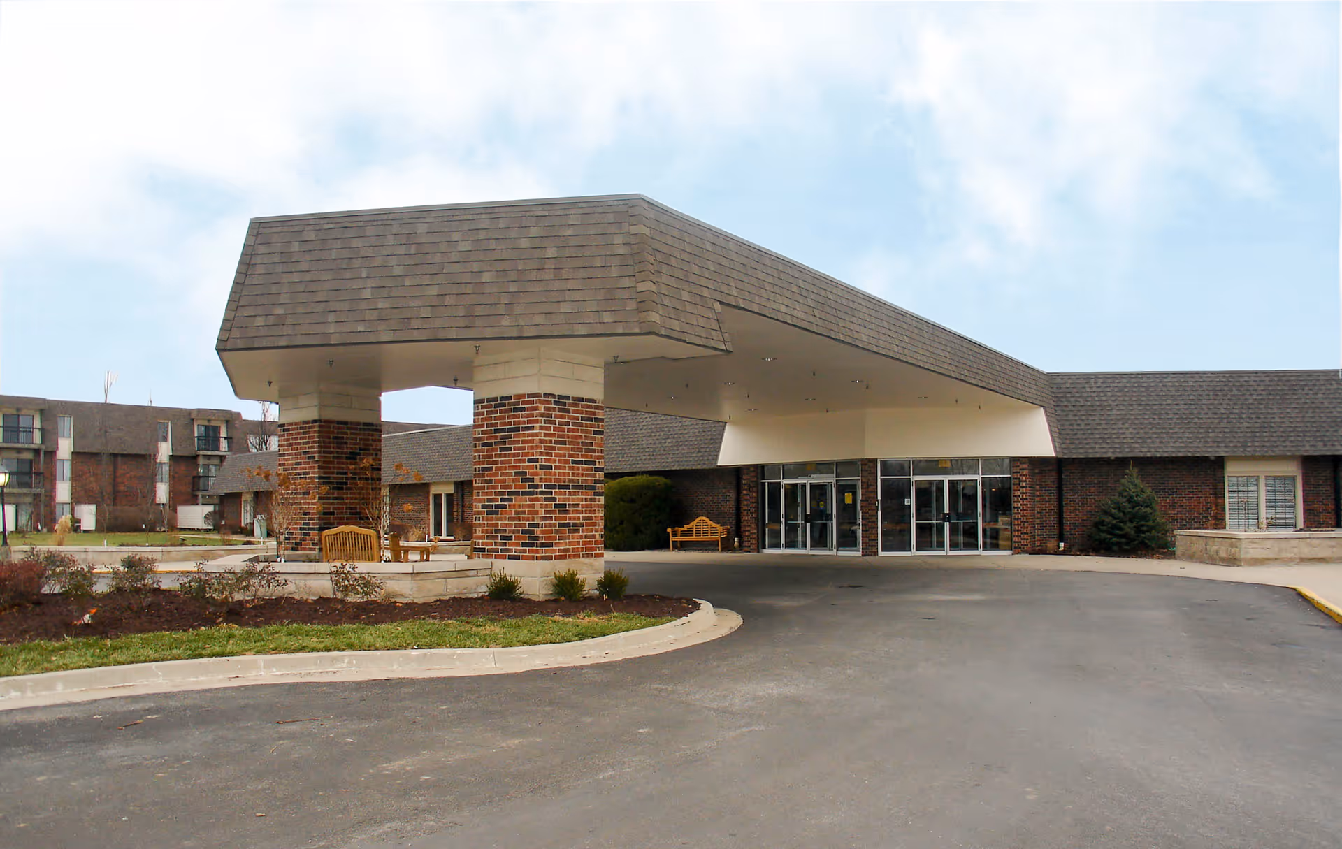 Front exterior view of Foxwood Springs Senior Living facility showing a covered entrance with brick pillars, glass double doors, benches, and a driveway leading up to the entrance.
