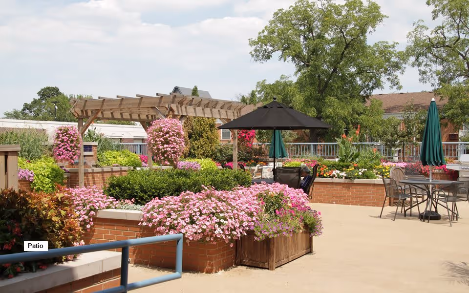 Outdoor patio area with brick planters filled with pink flowers and green shrubs, a wooden pergola, patio tables with umbrellas, and trees in the background under a partly cloudy sky.