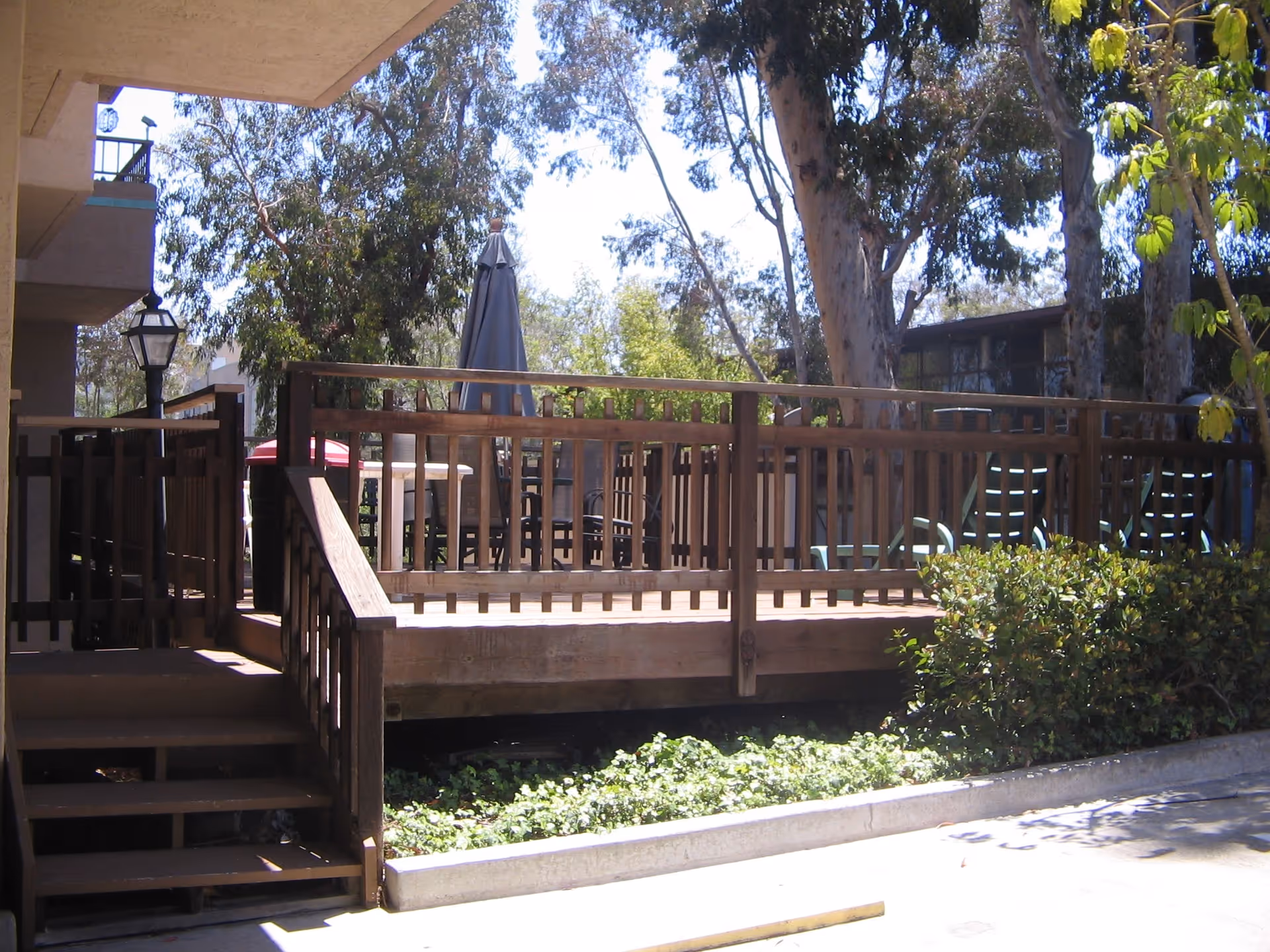 Outdoor wooden deck area with railings, patio furniture including chairs and tables with umbrellas, surrounded by trees and bushes under a clear sky.