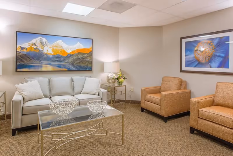 A cozy living room with a light gray sofa adorned with two white pillows, two brown armchairs, a glass coffee table with decorative bowls, two side tables with lamps, and two large framed artworks on the walls featuring a mountain landscape and a close-up of a blue flower.