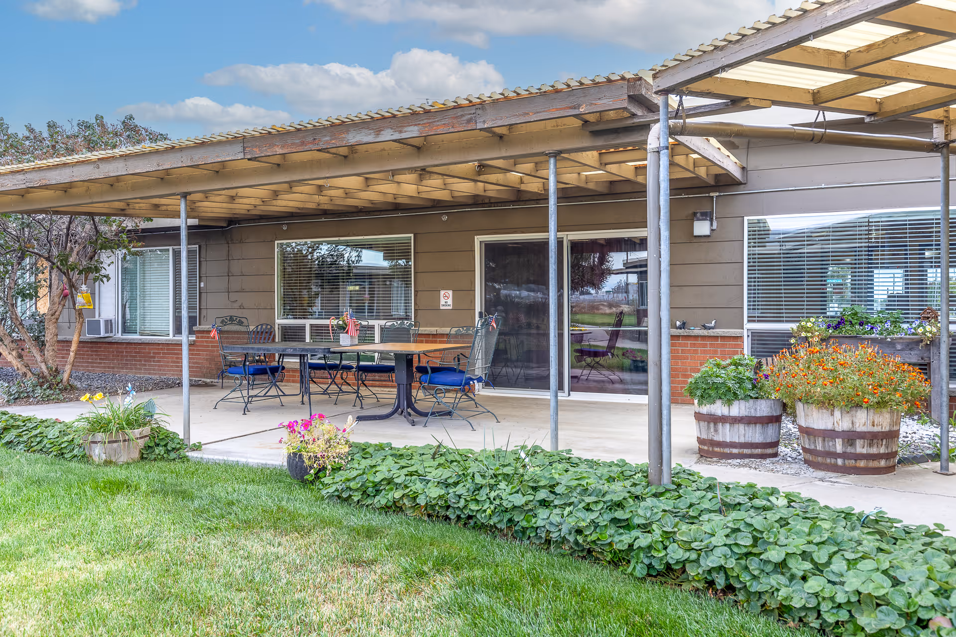 Covered outdoor patio with tables and chairs, potted flowers, and sliding glass doors on a single-story building.