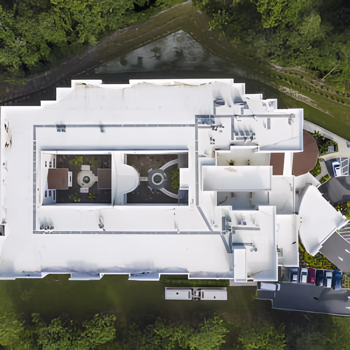 Aerial top-down view of a large white-roofed building surrounded by greenery and parking areas, showing the layout and structure of the facility.