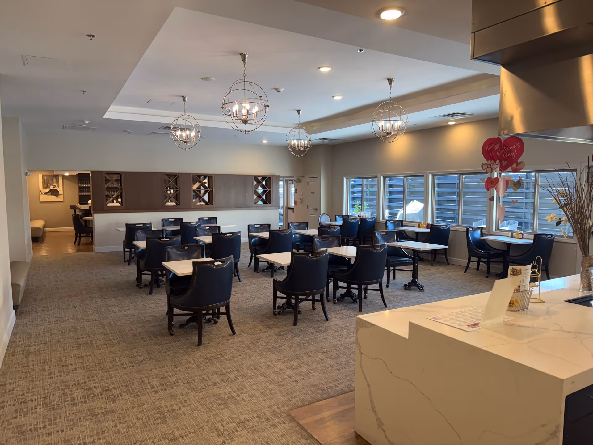 A spacious dining room in a senior living facility with multiple tables and black chairs arranged neatly. The room has modern spherical pendant lights hanging from the ceiling, large windows letting in natural light, and a white marble countertop in the foreground. There are Valentine's Day decorations hanging near the countertop.