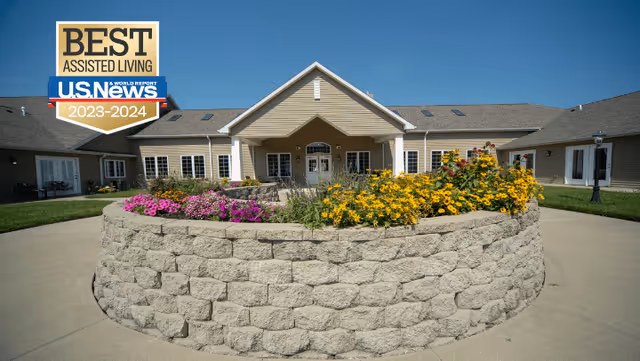 Exterior view of a single-story assisted living facility building with beige siding and a peaked roof. In front of the building is a circular stone planter filled with colorful flowers including yellow and pink blooms. The sky is clear and blue.