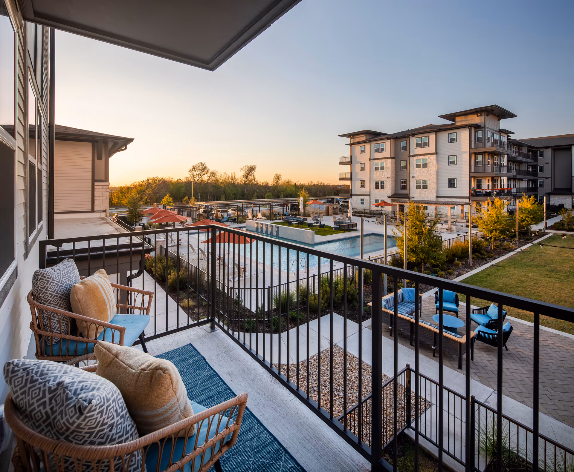 View from a balcony with two cushioned wicker chairs and a blue rug, overlooking a swimming pool area with lounge chairs, umbrellas, and a seating area with blue cushions. In the background, there is a multi-story residential building and trees under a clear sky at sunset.