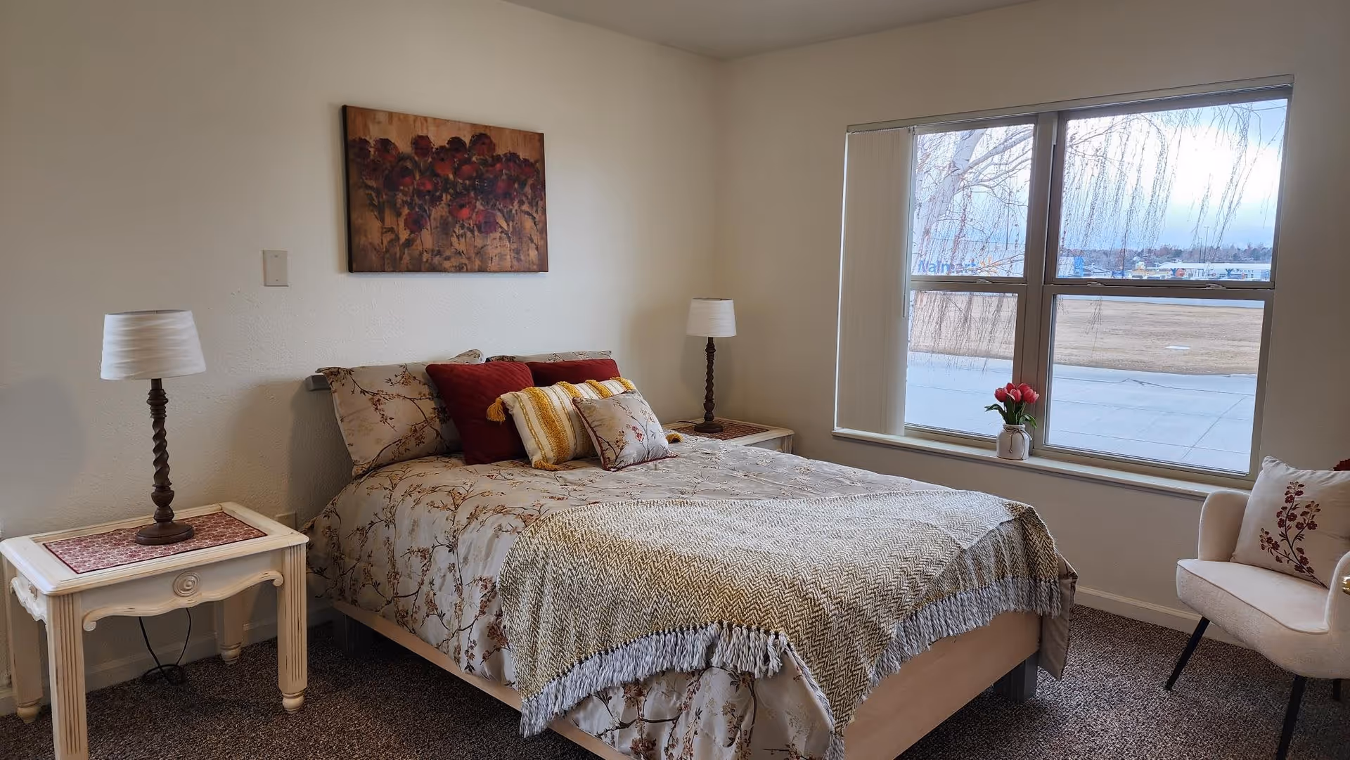 A cozy bedroom with a bed featuring floral bedding and multiple pillows. There are two bedside tables, each with a lamp. A painting of red flowers hangs above the bed. A large window lets in natural light, and a small vase with flowers sits on the windowsill. A cushioned chair with a decorative pillow is placed near the window.