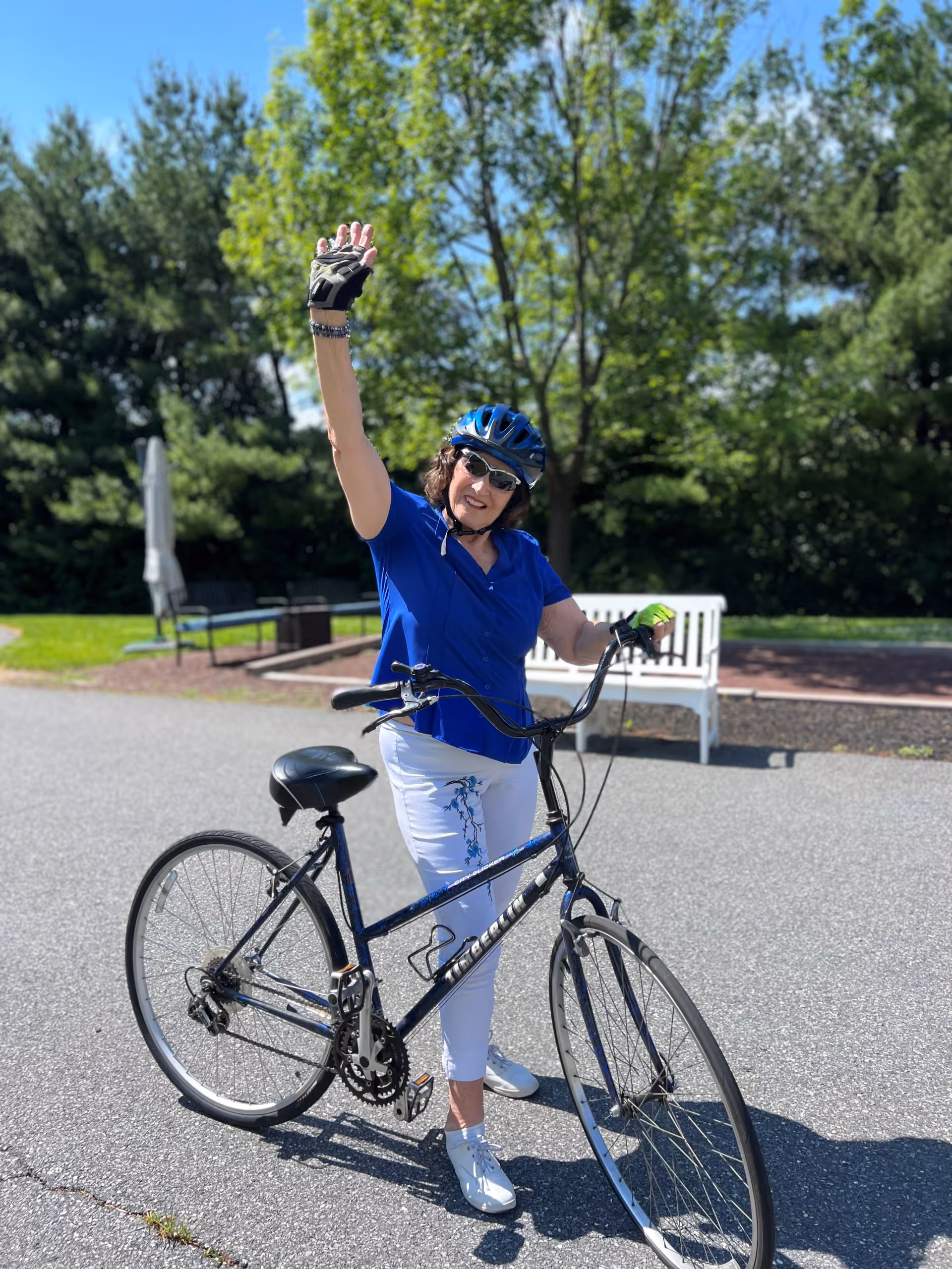 A woman wearing a blue helmet, sunglasses, a blue shirt, and white pants is standing outdoors on a paved surface with a black bicycle. She is smiling and waving with her right hand raised. Behind her, there are green trees, a white bench, and some outdoor furniture under a clear blue sky.