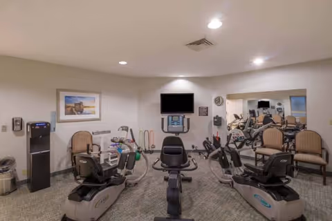 Interior view of a fitness room with exercise equipment including stationary bikes and elliptical machines. There are chairs along the wall, a water dispenser, a wall-mounted TV, and a large mirror reflecting the room.