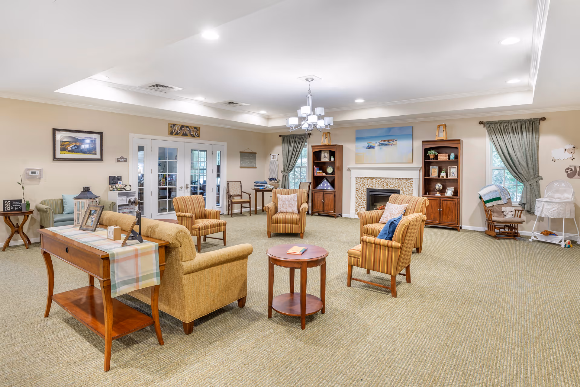 A spacious and well-lit living room area with beige walls and carpet. The room features several comfortable armchairs arranged around a small round wooden table. There is a fireplace with a decorative mantel and two wooden bookshelves on either side. The room has large windows with green curtains, a chandelier hanging from the ceiling, and a wooden console table with framed photos and decorative items. French doors lead to another room in the background.