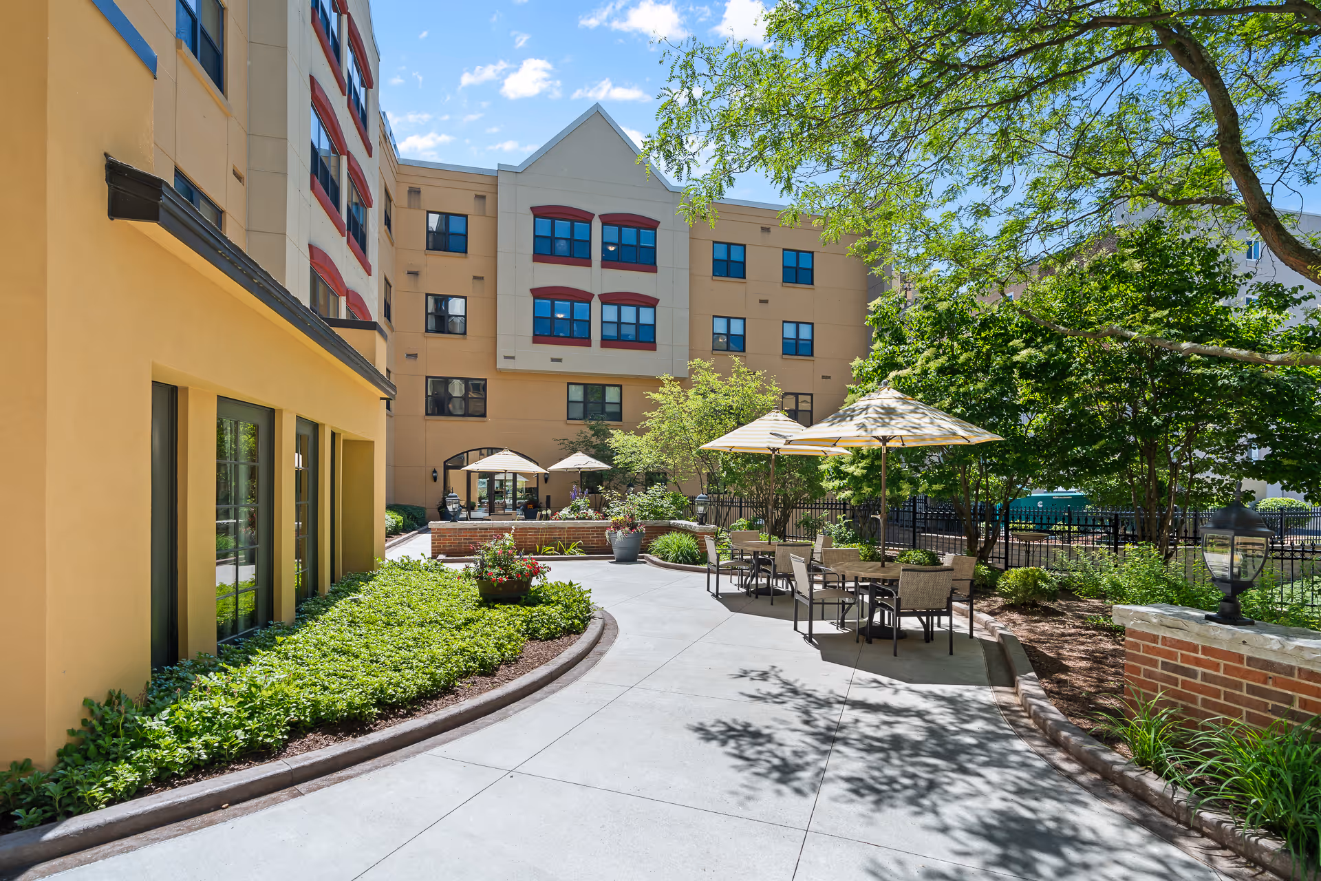 Outdoor patio area at Avalon Square with tables, chairs, and umbrellas surrounded by greenery and trees, adjacent to a multi-story building under a blue sky with some clouds.